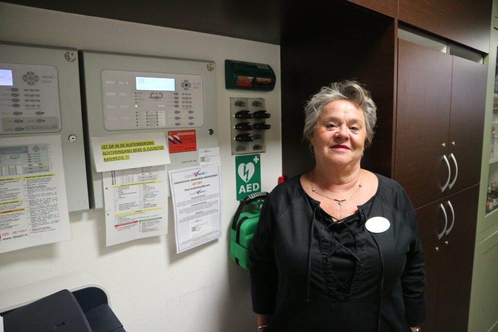 Caroline Paanakker staat naast de brandmeldcentrale en AED op een woonzorglocatie van DrieGasthuizenGroep.