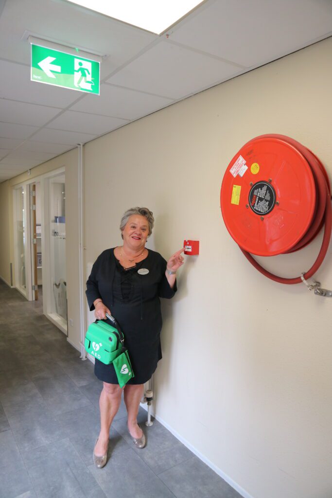 Caroline Paanakker wijst naar een handbrandmelder en houdt een AED vast.