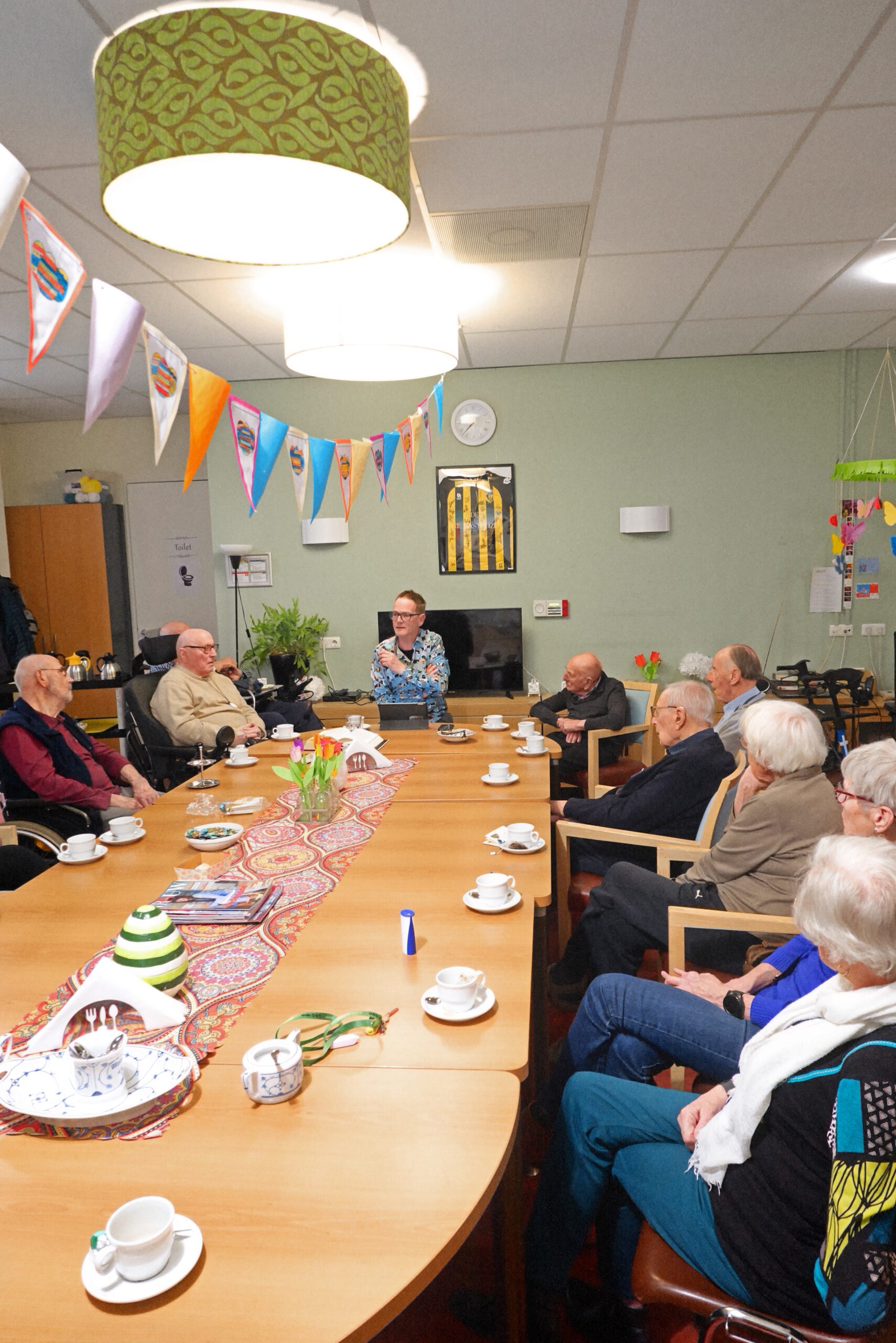 Bewoners zitten rond de tafel in zaal Hofstaete tijdens de wekelijkse voorleesavond op Insula Dei.